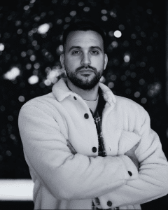 Retrato para sección Sobre mí: hombre con chaqueta blanca y brazos cruzados, fondo bokeh oscuro