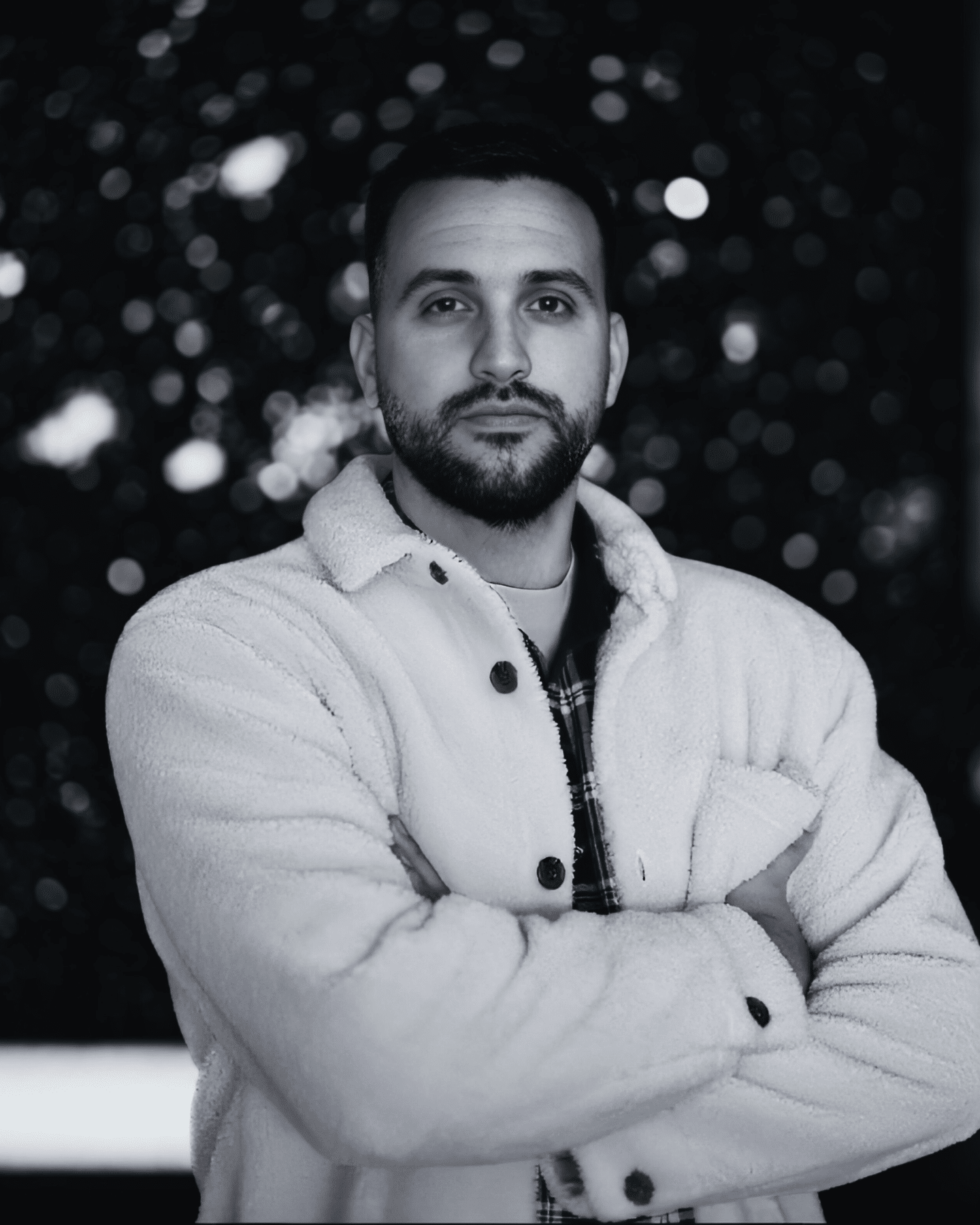 Retrato para sección Sobre mí: hombre con chaqueta blanca y brazos cruzados, fondo bokeh oscuro