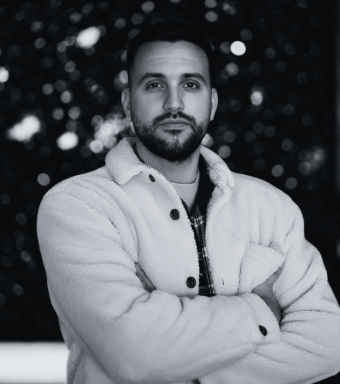 Retrato para sección Sobre mí: hombre con chaqueta blanca y brazos cruzados, fondo bokeh oscuro